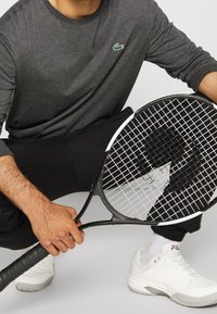 Šedé tričko s dlouhým rukávem a malým zeleným logem, černé kalhoty, drží černou teniskovou raketu, na nohou bílé sportovní boty, texturovaný úchop na rukojeti.