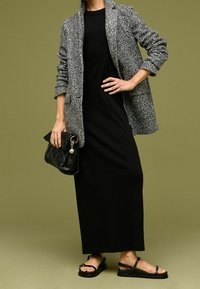 Blazer gris texturizado sobre un vestido largo negro, combinado con un bolso acolchado negro y sandalias negras. El fondo es de color verde oliva.