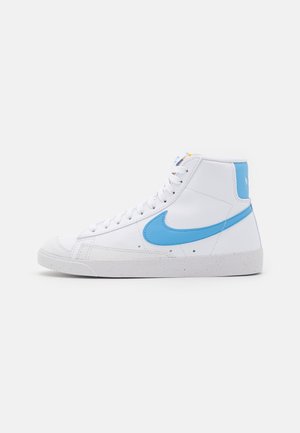 Biały sneaker typu high-top z jasnoniebieskim logo Nike i zakładką na pięcie, białymi sznurowadłami oraz nakrapianą białą podeszwą na jednolitym tle.