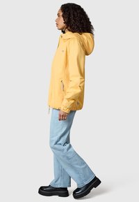 Donna con capelli ricci che indossa una giacca gialla con cappuccio, jeans azzurri e scarpe platform nere, in piedi di profilo su sfondo neutro.