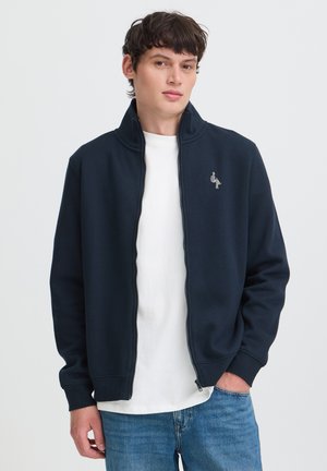 Giovane uomo con giacca blu navy con zip sopra una camicia bianca e jeans blu, in piedi con una mano in tasca.