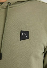 Oliwkowy hoodie o miękkiej fakturze, z małą czarną prostokątną naszywką z białym abstrakcyjnym wzorem oraz sznurkiem do regulacji.