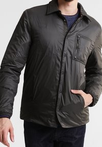 Chaqueta acolchada de color gris oscuro con un bolsillo en el pecho con cremallera, cuello de camisa, botones a presión y puños elásticos, hecha de material ligero.