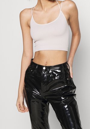 Kvinde iført en ribbet lysebeige cropped top med spaghetti stropper og skinnende sorte vinylbukser mod en ensfarvet baggrund.