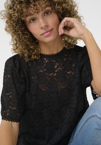 Black lace blouse featuring puffed sleeves, scalloped neckline, intricate floral patterns, and a textured mesh design. Worn with blue jeans.