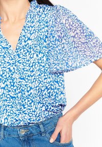 Una donna indossa una blusa blu con un motivo e maniche a sbuffo, abbinata a jeans di denim, con la mano appoggiata nella tasca.