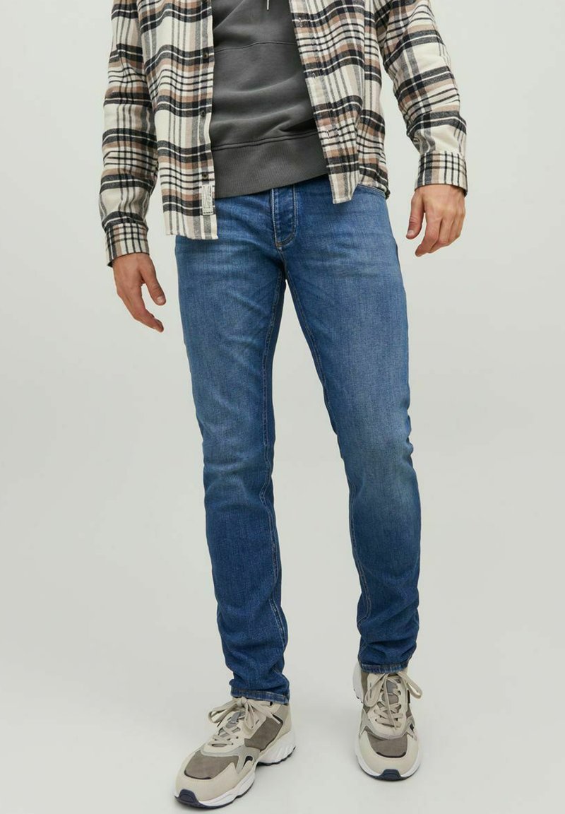 Vaqueros de mezclilla azul con un corte ajustado y un ligero estiramiento. Combinados con una sudadera gris y una camisa de franela a cuadros en tonos beige, marrón y negro.