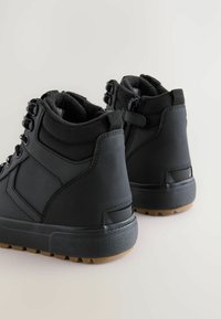 Schwarze Knöchelboots mit strukturiertem Stoff und Gummisohlen, ausgestattet mit Ösen für Schnürsenkel und einem gepolsterten Schaft, von hinten gezeigt.