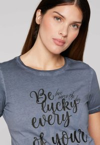 Blue cotton t-shirt with short sleeves, round neckline, and black text graphic reading "Be lucky every day" in varied fonts.