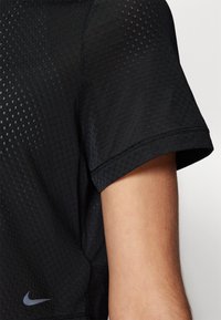 Melnā Nike sporta krekls ar teksturētu, elpojošu audumu ar maziem dimanta rakstiem, īsām piedurknēm un logotipu tuvu apakšmalai.