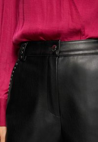 Sorte læderbukser med en glat tekstur, der har knaplukning og sølvnitter langs siden, parret med en magenta blouse.