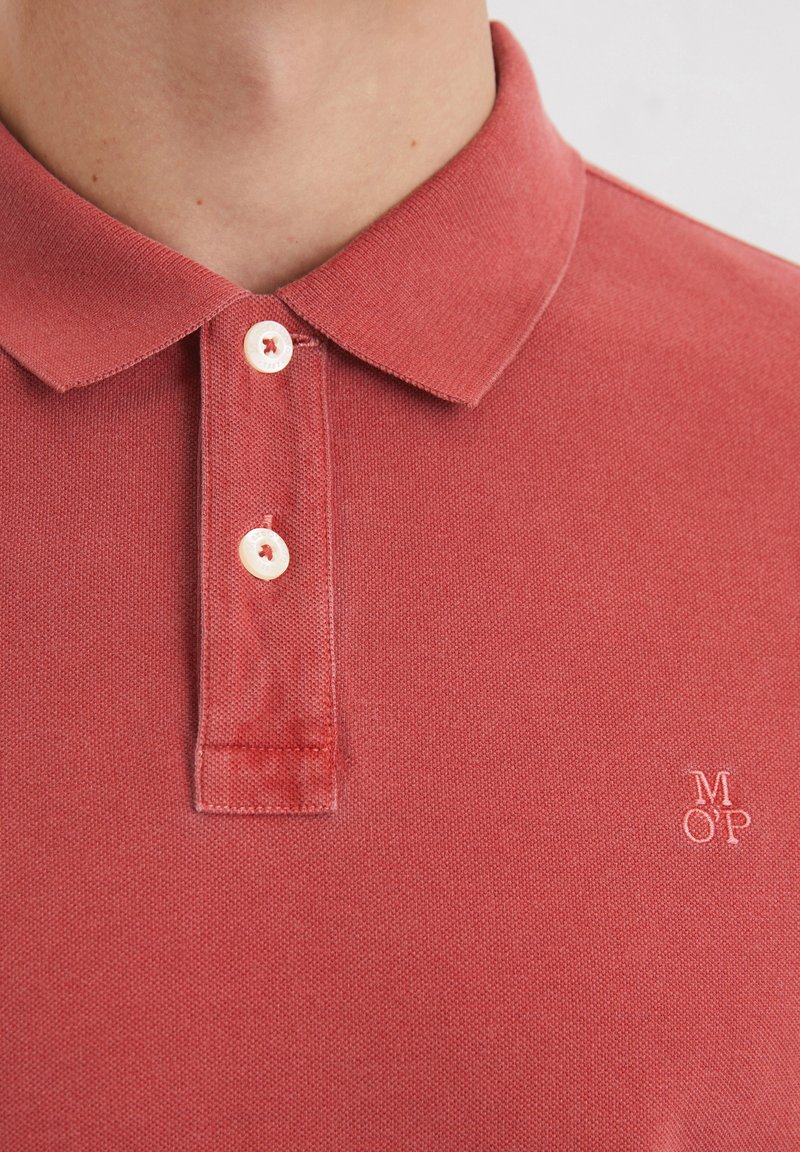 Polohemd aus weichem roten Stoff mit strukturiertem Finish, ausgestattet mit einem klassischen Kragen und drei weißen Knöpfen, bestickten Initialen auf der Brust.