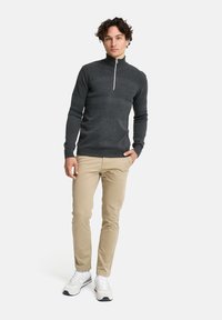 Harmaa vetoketjullinen neule, jossa on teksturoitu muotoilu, yhdistettynä beigeihin housuihin ja valkoisiin tennareihin. Malli seisoo yksinkertaisella taustalla.