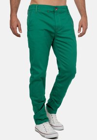 Pantaloni di cotone verdi con vestibilità slim. Presentano cuciture arancioni e chiusura con bottone. Indossati con sneakers bianche.
