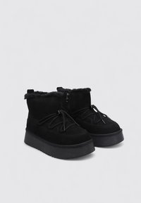 Botines de ante negro con forro acolchado, suela plana con plataforma y cordones decorativos. Cremallera lateral para un fácil acceso. Textura suave.