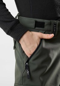 Zwarte ribgebreide lange mouw top gecombineerd met groene softshell broek met een ritszak en velc sluiting. Zwarte accentstiksels zichtbaar.