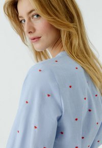 Hellblaue Bluse mit kurzen Ärmeln, verziert mit roten herzförmigen Stickereien, die über den Stoff verteilt sind. Glatter Stoff und lässiges Design.