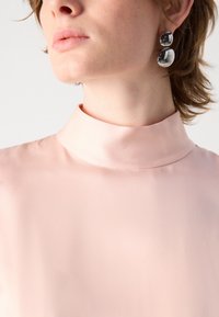 Blusa rosa a collo alto realizzata in tessuto liscio e lucido, con un colletto aderente. Sono visibili orecchini circolari in argento, che aggiungono un tocco di dettaglio.