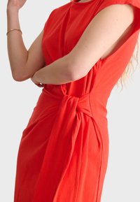 Robe rouge avec un détail de taille nouée, des manches courtes et un tissu doux, avec une silhouette ajustée mettant en valeur les courbes.