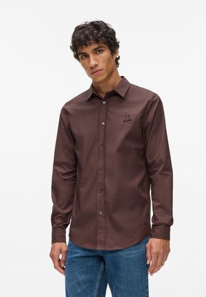 Camisa marrón de manga larga, abotonada y con un logo sutil, que presenta una textura suave y un corte ajustado. Combinada con jeans azules.