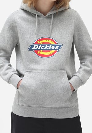 Osoba nosící světle šedou mikinu s kapucí a přední kapsou, s barevným logem Dickies na hrudi, ruce zasunuté v kapse.