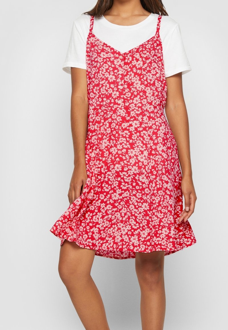 Femme portant une chemise blanche à manches courtes sous une robe sans manches rouge à fleurs avec des boutons sur le devant, se tenant devant un fond uni.