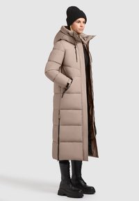 Hosszú bézs színű steppelt télikabát kapucnival, oldalán cipzárokkal és barna béléssel, fekete fűzős csizmával kombinálva.
