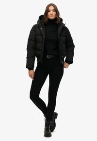 Chaqueta acolchada negra con capucha, diseño acolchado, cierre de cremallera, llevada sobre un cuello alto negro y jeans skinny negros, combinada con botas negras de cordones.