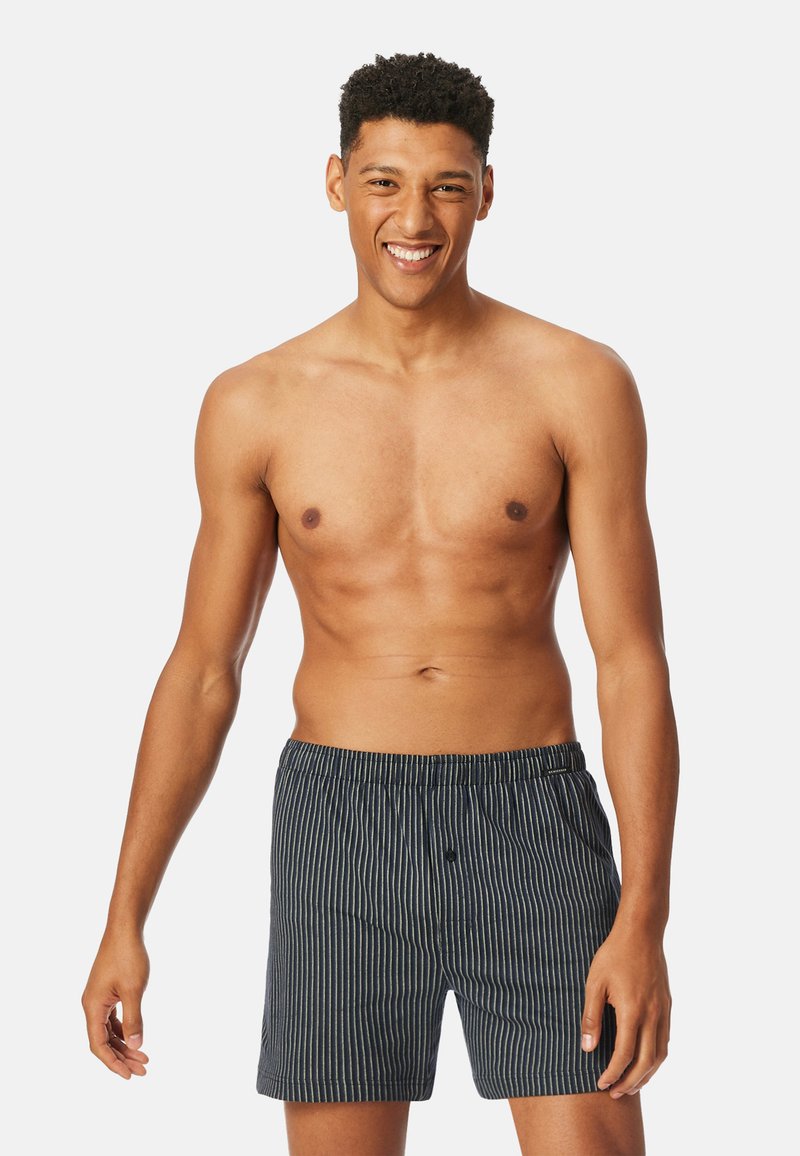 Marine- og hvitstripete bomullsboxere med elastisk midjebånd og en liten frontknapp. Modellen står med et nøytralt uttrykk og bar torso.