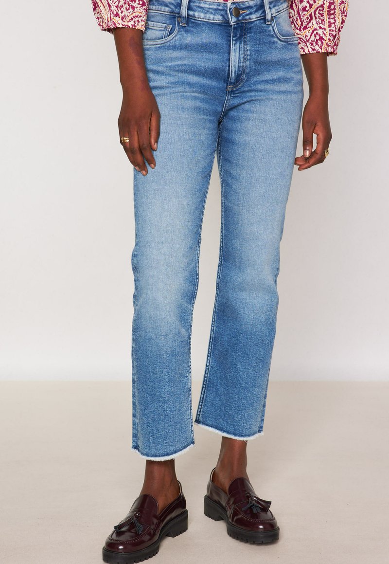 Jeans a gamba dritta di colore azzurro chiaro con orlo grezzo, dotati di cinque tasche, chiusura con zip e una texture denim classica. Abbinati a mocassini scuri.