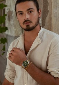 Silberne und goldene Uhr mit grünem Ziffernblatt, schwarzen Akzenten und leuchtenden Markierungen, kombiniert mit einem goldenen Kettenarmband, getragen zu einem beigen Leinenhemd.