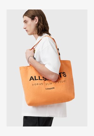 Borsa a tracolla in tela arancione con testo nero, che presenta la parola "ALLSAINTS" e "LONDON", con lunghe tracolle. Forma rettangolare semplice.