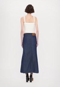 Witte gehaakte crop top met brede bandjes, gecombineerd met een lange blauwe denimrok met achterzakken en een bruine rechthoekige label.