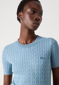 Maglione lavorato a maglia blu chiaro con un motivo a treccia, maniche corte e collo a costine. Presenta un piccolo logo ricamato sul lato sinistro del petto.
