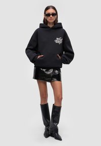 Czarny oversized hoodie z kieszenią z przodu i logo, połączony z błyszcząco czarną mini spódnicą i wysokimi czarnymi butami, na jednolitym szarym tle.