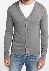 Cardigan grigio lavorato a maglia con scollo a V, maniche lunghe, bottoni frontali e polsini e orlo a coste; indossato sopra una maglietta bianca e jeans in denim.
