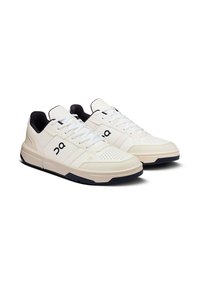 Baskets de sport blanches avec un dessus en cuir texturé et en suede, comportant des perforations, des accents noirs et une semelle rembourrée.