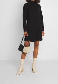 Robe pull noire avec un col haut, des manches longues et une texture côtelée. Bottines à talon bloc beiges claires et un sac à épaule bicolore.