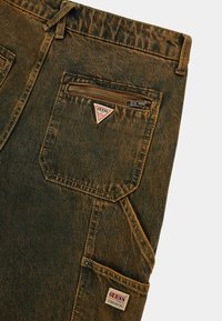 Denim cargobroek in een vervaagde bruintint met een gestructureerde afwerking. Kenmerkt zichtbare stiksels, twee zakken en een driehoekige logo patch.