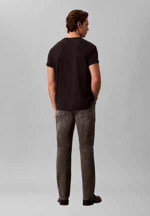 Mand stående med ryggen til, iført sort t-shirt med opkørte ærmer, falmede grå jeans og sorte sko mod en ensfarvet grå baggrund.