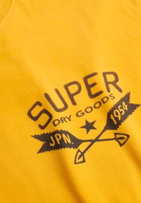 Gul bomulds-T-shirt med et sort grafisk tryk med teksten "SUPER DRY GOODS," en stjerne og pile, der fremhæver tekstur og brandingdetaljer.
