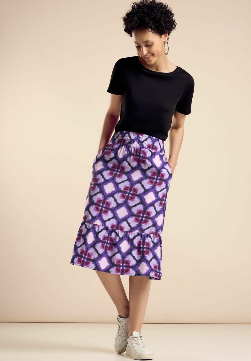 Falda morada con estampado, con un dobladillo con volantes y cintura elástica, combinada con una camiseta negra de mangas cortas y zapatillas blancas.