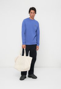 Blå hettegenser med rund hals og gult logo, sammen med svarte cargo-bukser. Holder en kremfarget tote bag med preget logo. Svarte joggesko.