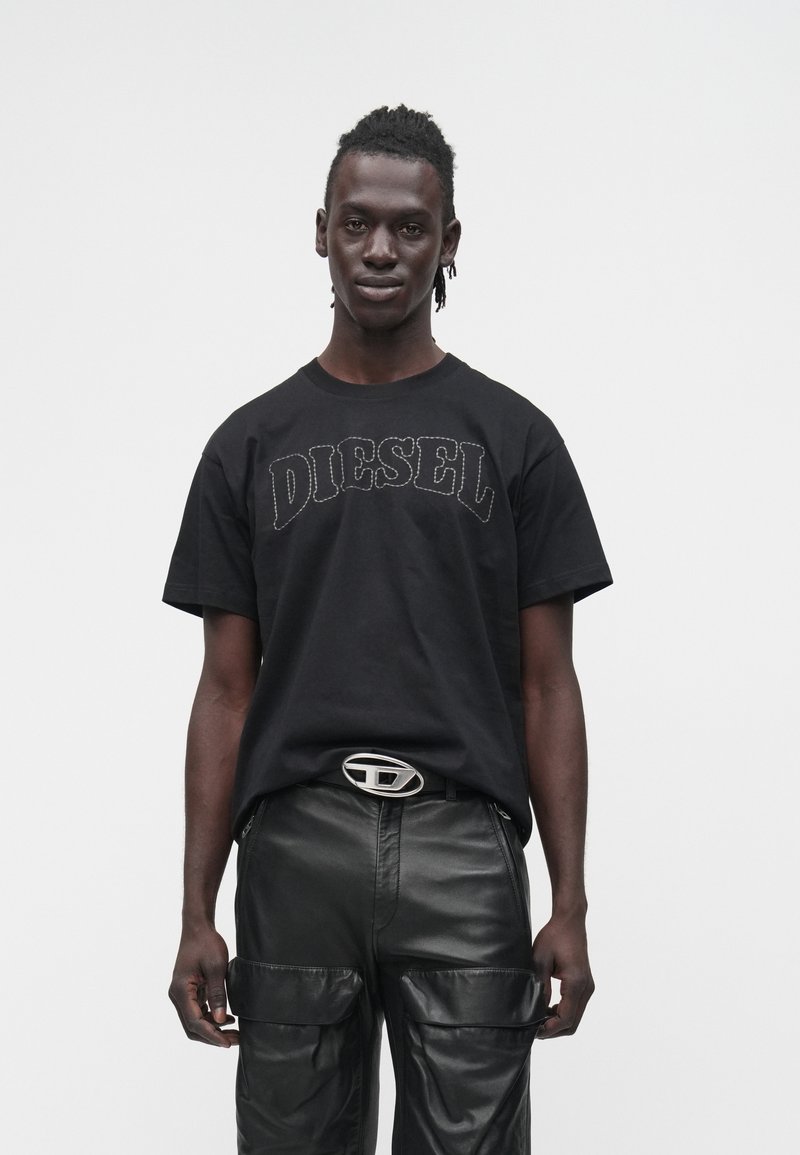 Mužský model v čiernom tričku Diesel s prešívaním, čiernych kožených nohaviciach s veľkými vreckami a opasku so striebornou prackou s logom Diesel.