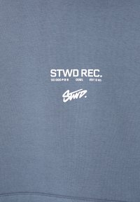 Sudadera azul claro con textura suave. Presenta texto impreso en blanco: "STWD REC. 00 000 P8 B 0091 EST 091" y un logo de firma en blanco.