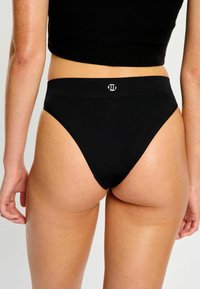 Femme portant un bas de bikini noir taille haute avec un logo sur la ceinture, vue de dos contre un fond blanc simple.