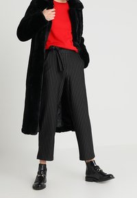 Manteau en fausse fourrure noire, pull en maille rouge, pantalon rayé noir, et bottines noires à boucle. Le pantalon est orné d'un détail de ceinture à nouer.