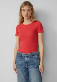 Czerwona bluzka na krótki rękaw z okrągłym dekoltem, w zestawie z jasnoniebieskimi dżinsami z wysokim stanem. Materiał ma gładką teksturę.