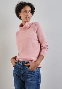 Rosa Strickpullover mit hohem, geknöpftem Kragen und strukturierten Schulterakzenten. Kombiniert mit blauen Jeans, die eine entspannte Passform zeigen.