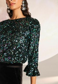 Top in paillettes nere con maniche a tre quarti e polsini arricciati, caratterizzato da paillettes riflettenti multicolori per un tocco di texture e brillantezza.